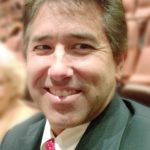 A smiling Darren Medeiros with short brown hair, wearing a black suit and a white shirt with a red patterned tie. He appears content and is seated in what looks like an auditorium, with blurred rows of seats in the background.