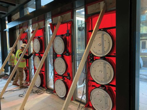 Two construction workers in safety gear are adjusting equipment on a wall where large circular devices are mounted, surrounded by red barriers. The area is still under construction, with visible wires and scaffolding.