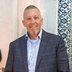 Edward Owens with short hair and a trimmed beard stands smiling, wearing a gray plaid blazer over a light blue shirt. He is positioned in front of a textured wall featuring colorful decorative tiles, creating a contrast between the modern attire and the traditional background.