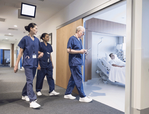 In a brightly lit hospital corridor, three healthcare professionals—two women and one man—walk towards a patient room where a bedridden patient is visible. The first woman, with her hair tied back, and the second woman, in scrubs, walk alongside a male colleague who is looking at a tablet. The atmosphere suggests a busy healthcare environment.