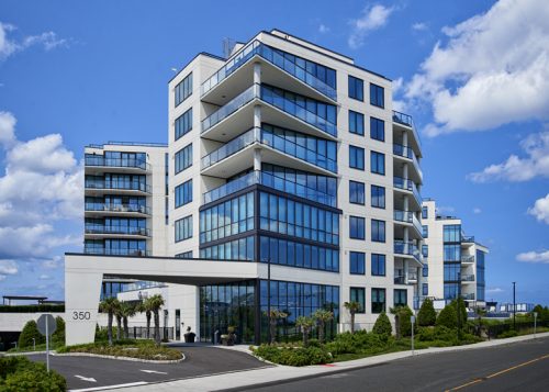 A contemporary residential building features multiple stories and large glass balconies. The building has a modern architectural design, with a wide entrance and landscaped greenery at the base. The sky is blue with a few fluffy clouds, creating a pleasant atmosphere.