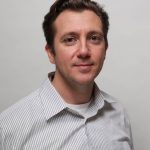 Michael Sellitto in a close-up portrait, smiling gently. He has short, dark hair and a slight stubble. He is wearing a white shirt with a striped gray and white button-up shirt. The background is a soft, light gray, emphasizing his features.