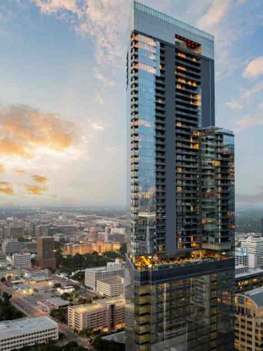 A modern skyscraper features a sleek glass facade, reflecting the colors of a sunset. The building stands tall amidst a cityscape, showcasing various architectural styles below. Warm lights glow from the windows, suggesting a vibrant atmosphere.