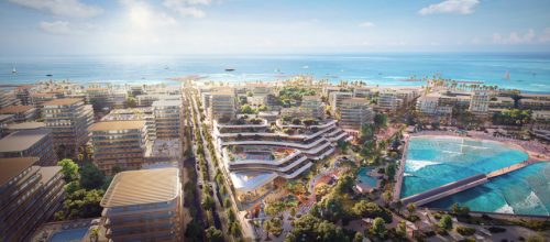 A vibrant coastal development featuring modern mid-rise buildings surrounded by lush greenery and pathways. The central area includes unique, wave-like architectural structures and recreational spaces. A large artificial lagoon with turquoise water and a wave pool is visible, alongside a sandy beach and the expansive ocean in the background, dotted with boats and sailboats under a sunny sky.