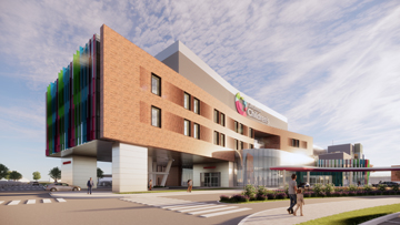 A modern hospital building featuring a multi-colored façade and large windows. The entrance is spacious, with an inviting design. Pedestrians are visible in the foreground, and a clear blue sky enhances the setting.