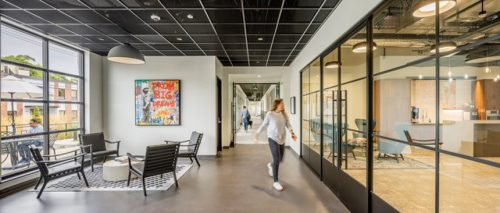 A contemporary office corridor is depicted, featuring a cozy seating area with black chairs and a round table. A large, colorful artwork titled "Dream Big Dreams" is hung on the wall. The corridor leads to glass-walled meeting rooms and a kitchen area, with windows allowing natural light and a view of an outdoor space. A person is walking through the corridor, and another individual can be seen in the background.