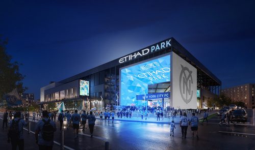 A night view of Etihad Park, home of New York City FC, filled with fans gathering at the entrance. Bright blue lights illuminate the stadium's façade, showcasing the text "Welcome to Etihad Park" and the New York City FC logo. The atmosphere is vibrant with fans wearing team colors and waving flags.