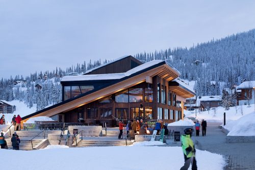 A modern ski lodge features large glass windows and a sloped roof, set in a snowy landscape. Groups of people are gathered outside, some sitting on benches, while others walk by or interact. The lodge's warm interior glow contrasts with the cold, snowy surroundings.