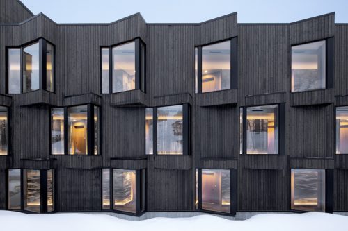 A contemporary wooden building features a façade with irregularly shaped, protruding windows. The structure is set against a snowy landscape, with soft lighting visible through the windows, suggesting a warm interior atmosphere.