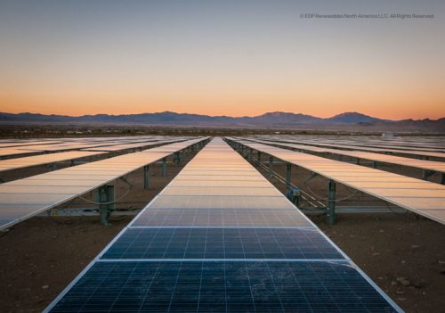 The image shows a vast solar panel array stretching into the horizon, with rows of reflective panels aligned neatly on the ground. The sky is painted with warm colors as the sun sets behind distant mountains, creating a serene atmosphere. This scene emphasizes renewable energy and sustainable practices.