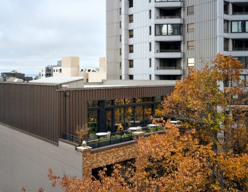 A modern building features a flat brown roof and expansive glass windows on one side. Below, a small terrace with several white tables and chairs is visible, framed by trees with autumn-colored leaves. In the background, a taller building stands beside the structure.