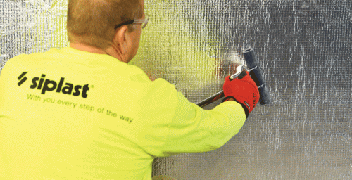 A worker wearing a bright yellow long-sleeve shirt with the "Siplast" logo is using a roller to apply reflective insulation material to a surface. The worker is wearing red gloves and has safety glasses on, ensuring protection while working.