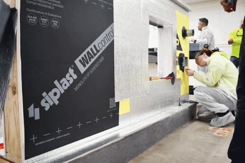 In a construction training environment, a person in protective clothing kneels to apply a product to a wall. The wall features a black panel labeled “siplast WALLcontrol” and yellow markings indicating points of interest. Other attendees are positioned in the background, engaging in the workshop.