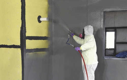 A person wearing a protective suit and mask is spray painting a wall. The wall is primarily gray with a section of yellow, and a pipe is visible on the surface. There is a window in the background, highlighting the room's unfinished state.