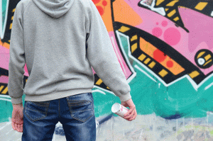 A person wearing a gray hoodie and blue jeans stands with their back to the viewer, holding a cup in their right hand. The background features a vibrant graffiti wall with pink, green, and black colors, adding an urban artistic vibe to the scene.