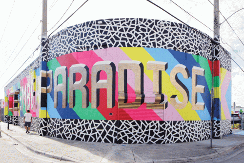 A vibrant mural wraps around a building, featuring the bold word "PARADISE" in large, 3D letters. The mural comprises bright colors like pink, blue, and green, with abstract geometric patterns throughout. A person in a white shirt walks along the sidewalk in front of the mural.