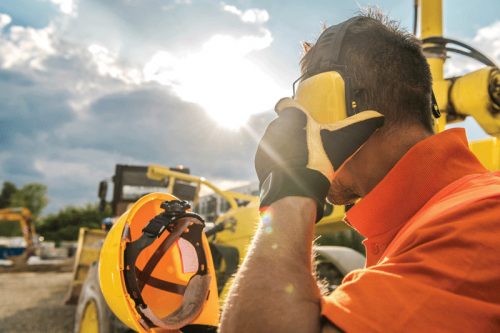 A construction worker, wearing an orange safety shirt and ear protection, is holding a phone to his ear while standing on a construction site. In the background, yellow machinery is visible against a dramatic sky with sunlight streaming through clouds.