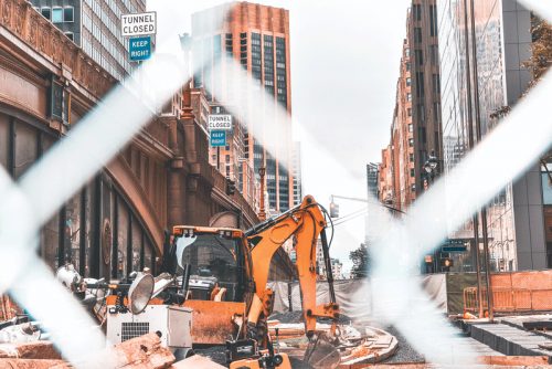 Urban construction scene with an excavator and 'Tunnel Closed' signs in a city surrounded by tall buildings, seen through a chain-link fence.