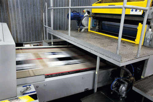 A large industrial printing machine features a conveyor belt below a metallic platform. The machine has a bright yellow roller and various control panels. The conveyor transports printed materials, showing motion blur, indicating operation in a factory setting.