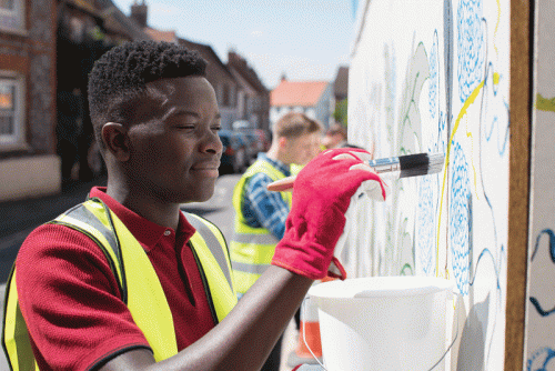 A young man wearing a red shirt and reflective vest smiles as he paints a mural on a wall with a brush. He holds the brush in his right hand, while his left hand rests against the wall. In the background, a person in a blue and green shirt is also painting. There are residential buildings and a clear blue sky visible.