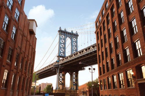 View of the Manhattan Bridge framed by red brick buildings in New York City, with a bright blue sky in the background.