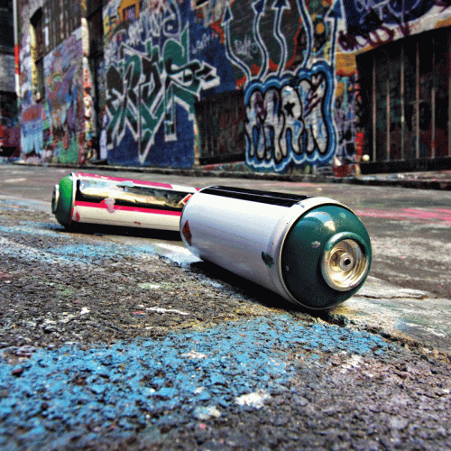 Two used spray paint cans are resting on a textured street surface, surrounded by vibrant graffiti on brick walls in the background.