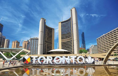 A vibrant scene of Toronto's city hall, showcasing two iconic curved towers. In the foreground, a large "TORONTO" sign stands by a reflecting pool, surrounded by people enjoying the public space. Clear blue skies enhance the lively atmosphere.