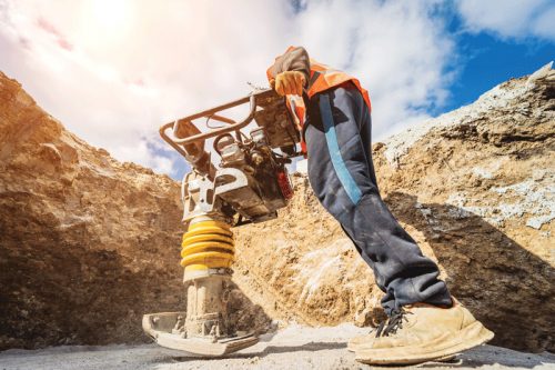 A construction worker is using a compacting machine in a trench. The worker wears an orange safety vest and dark pants, with the machine featuring a yellow spring beneath. The background shows soil and a clear blue sky with some clouds.