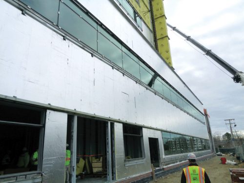 A construction site featuring workers installing large glass windows on the silver, reflective exterior of a building. The top of the building is being worked on using a crane. The sky is overcast, indicating the scene may be taking place in cooler weather.