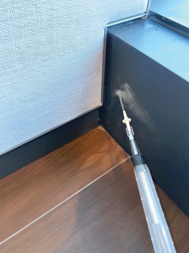 A syringe is positioned in the corner where the floor meets the wall, with a white substance visibly being dispensed. The background features a textured gray surface next to a black wall edge, and the wooden floor reflects light, enhancing visibility.