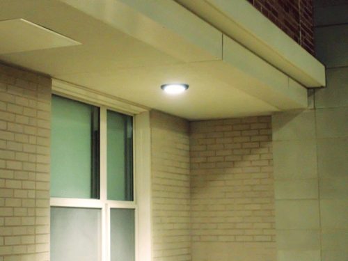 A building corner featuring a modern design. There is a bright ceiling light mounted above a large window, which has multiple panes and light-colored frames. The wall is made of light-colored bricks, creating a contrast with the dark night sky.