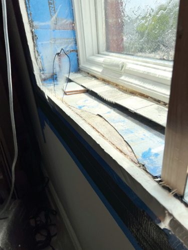 A close-up view of a damaged window frame featuring a section of cracked glass. The frame shows exposed insulation, with blue painter's tape applied around the edges. The window is partially foggy, and the surrounding wall is visible, indicating ongoing construction or repair.