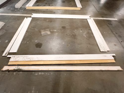 A collection of wooden planks lies on a concrete floor, arranged to form a square. The planks include several white-painted pieces and two unfinished wood boards. The floor shows visible marks and stains, indicating wear over time.