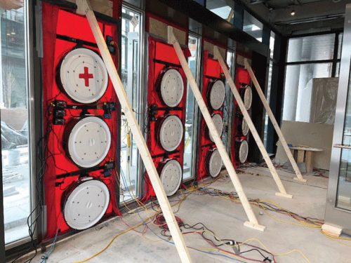 Inside a construction site, several red walls display circular equipment featuring a plus sign. Wooden beams support the structures, and messy electrical wires are visible on the floor, indicative of ongoing work. Large windows in the background allow light to enter.