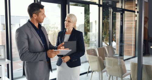 A man and a woman are conversing in a contemporary office environment. The man holds a tablet and gestures while speaking, and the woman listens attentively, holding a folder. Large windows in the background let in natural light, with stylish chairs arranged nearby.