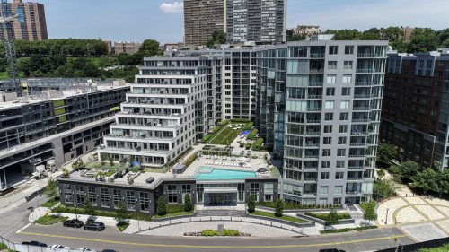 Aerial view of a modern apartment complex featuring multiple buildings with large windows. The foreground shows a landscaped courtyard, while the background reveals taller residential buildings.