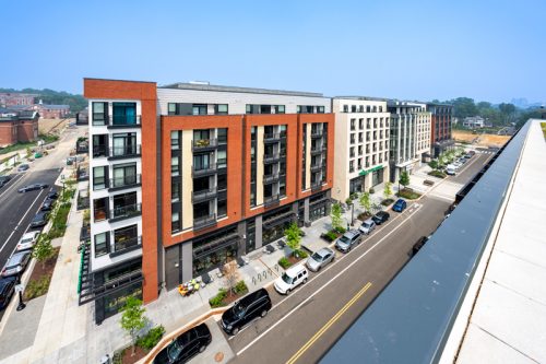 Modern urban apartment complex with green areas, street parking, and multiple balconies. Bright, sunny day with a clear sky, conveying a lively atmosphere.