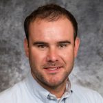 Chris Kann with short hair and a beard wears a light blue button-up shirt. The background is a textured gray, conveying a professional tone.