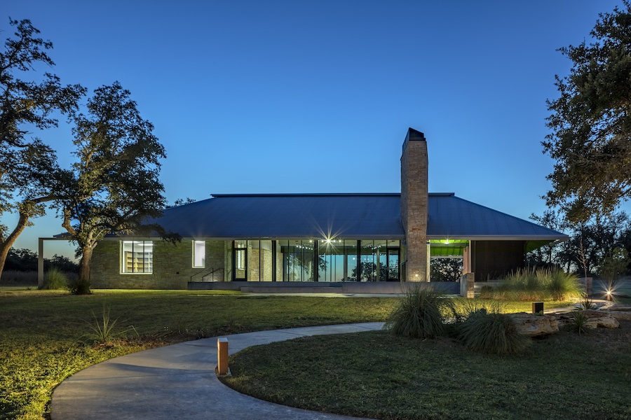 Modern architectural home at dusk, featuring large glass windows, stone chimney, and landscaped surroundings.