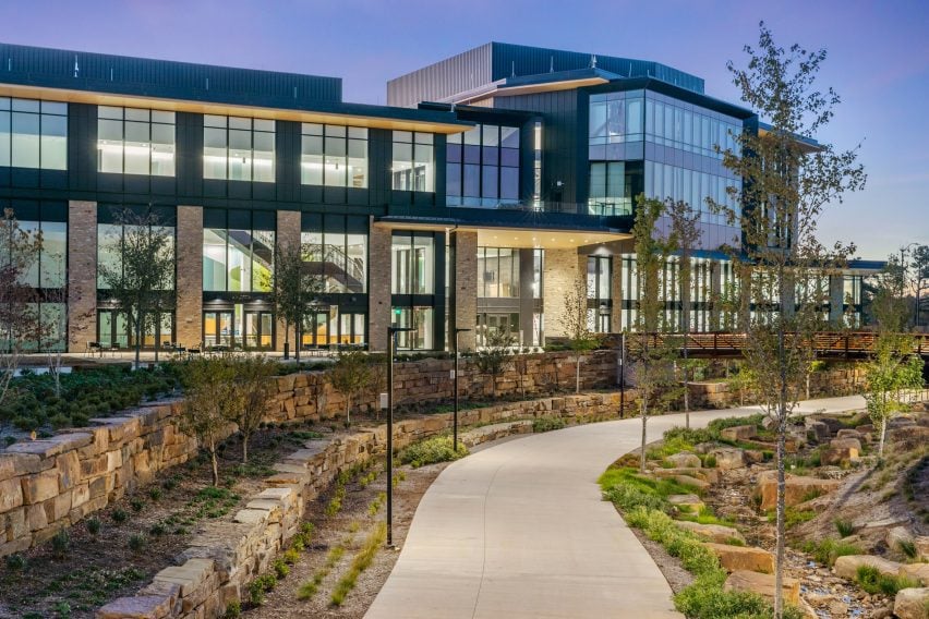 Modern building with large windows surrounded by landscaped paths and greenery at dusk.