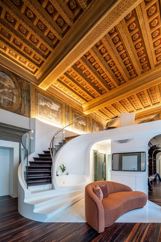 Luxurious interior with ornate coffered ceiling, modern white spiral staircase, and curved brown sofa. Elegant fusion of classical and contemporary design.