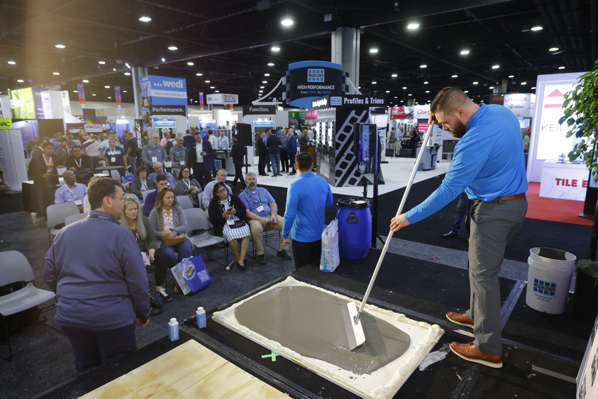 A man demonstrates floor leveling with a trowel in front of a seated audience at a busy trade show. Signs and product booths fill the background.