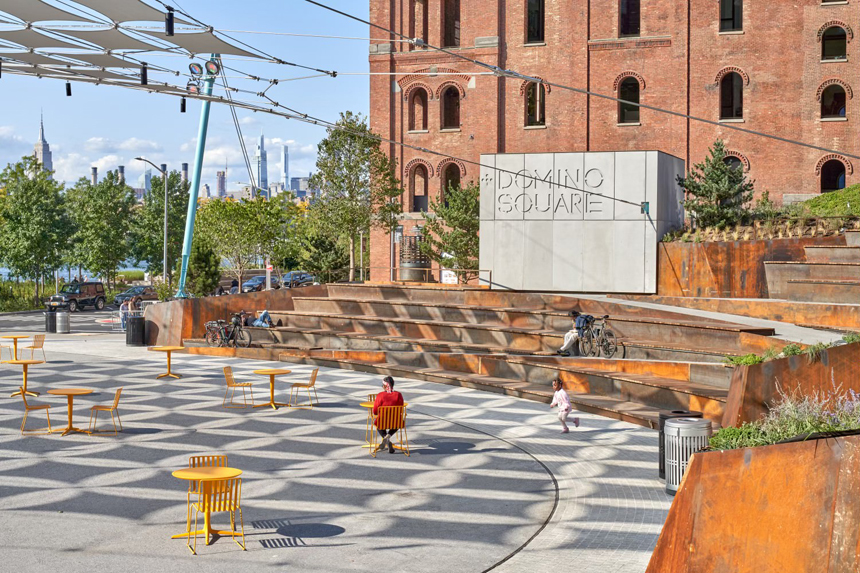 Domino Square in Brooklyn, featuring yellow outdoor tables and chairs, tiered rust-colored seating, and a modern canopy casting geometric shadows, with a historic brick building and the Manhattan skyline in the background.