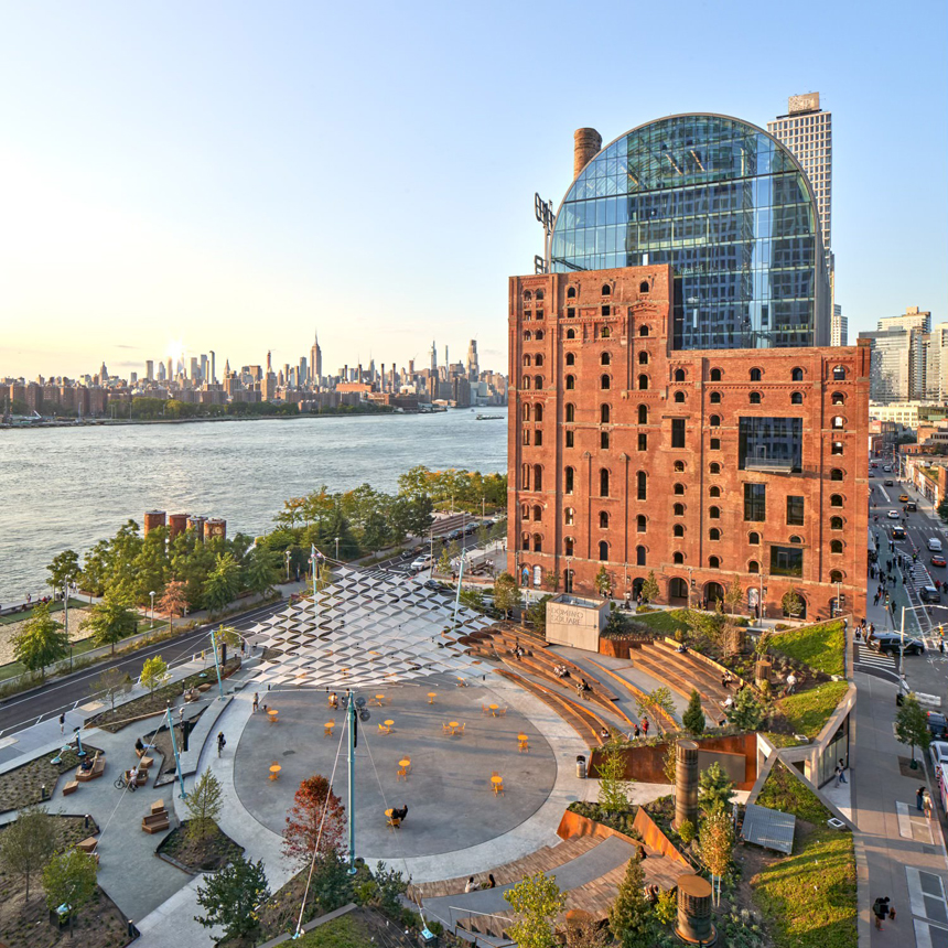 Aerial view of Domino Square in Brooklyn, showcasing a restored red brick refinery building with a modern glass addition, tiered seating, and a public plaza with yellow tables and chairs.