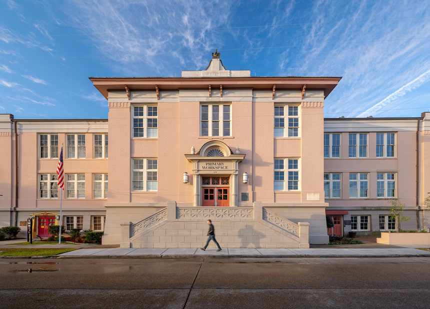 Historic pink school building with a central entrance labeled 'Primary Workspace,' featuring symmetrical architecture, large windows, and a decorative rooftop sculpture, with a person walking by in the foreground