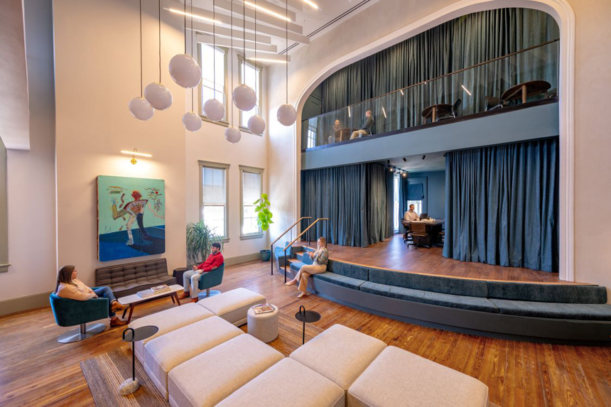 Modern coworking space with high ceilings, stylish seating, a small stage area with tiered seating, and an upper-level workspace behind dark curtains, featuring people working and relaxing.